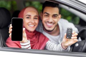 contento islámico esposa en hijab y marido espectáculo llave y inteligente teléfono con blanco pantalla en abierto ventana en auto, al aire libre en verano. comprando nuevo auto, solicitud, mapa y moderno tecnología, vida seguro foto