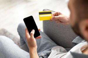 Ordering goods online. Unrecognizable young guy with credit card and smartphone shopping on web at home, mockup. Closeup of millennial man purchasing products in internet, using new mobile app photo