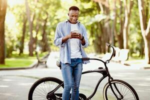 Funky black guy with bicycle sending communicating online on smartphone at park. Handsome African American man checking mail, browsing web, sending message or posting in social media outside photo
