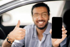 Happy arab man driver holding cellphone with blank screen, mockup. Closeup of mobile phone with empty display in smiling middle-eastern guy hand, using mobile app for navigation, copy space photo