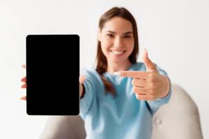 Joyful millennial woman pointing at tablet with blank screen for mockup, promoting new app, offering space for website or advertisement, selective focus. Touch pad display template photo