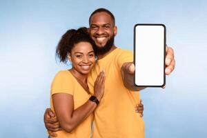 New awesome mobile app. African American couple presenting smartphone with mockup, promoting application or website, advertising your product or service, blue studio background. Selective focus photo