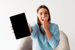 Shocked young woman showing tablet computer with black blank screen for mockup, covering mouth in surprise, advertising new app, offering empty space for website, white background. Selective focus photo