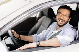 Cheerful long-haired arab guy with glasses driving right wheel auto, looking through open window and smiling at camera, happy young middle-eastern man enjoying car ride, copy space photo