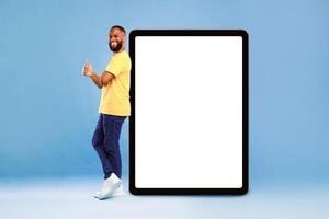 Positive African American guy showing thumb up gesture next to huge digital tablet on blue studio background, empty space for app design on screen. Touch pad with website or advertisement mockup photo