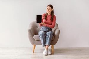 Smiling woman in casual wear showing tablet with empty screen, sitting in armchair against white wall, mockup for application or website. Young lady presenting touch pad with space for template photo