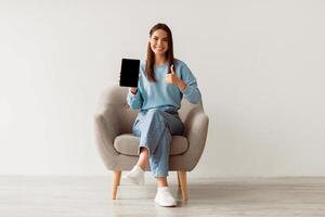 Smiling millennial woman showing empty touch pad, gesturing thumb up, sitting in armchair against white wall, mockup for website design on tablet screen. Online ad concept photo