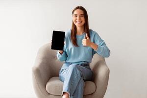 Charming millennial woman showing tablet pc with empty screen, gesturing thumb up in cozy armchair on white studio background, mockup for website or new app design. Touch pad display template photo
