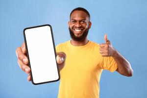 New cool mobile app. Funky African American guy demonstrating cellphone with empty screen, showing thumb up gesture, recommending website, offering space for advertisement on blue background, mockup photo