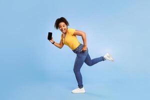 Hurry, big online sale. Happy young African American woman with empty smartphone running on blue studio background, mockup for web store website or mobile app. Space for your advertisement template photo