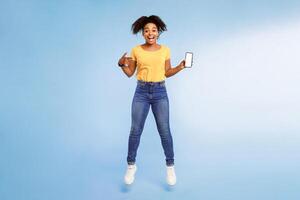 Cool mobile app. Excited African American woman pointing at smartphone with blank screen and jumping over blue studio background. Free space for mockup, advertisement, website or application design photo