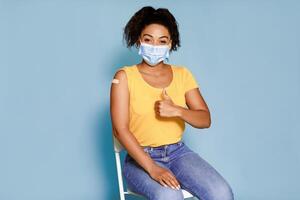 Portrait of young African American woman with band aid after flu vaccination showing thumb up, approving of flu immunization over blue studio background. Vaccine saves lives concept photo