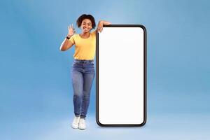 New awesome mobile app. Positive African American lady posing near huge cellphone with empty screen, showing okay gesture, recommending website, presenting space for advertisement on blue photo