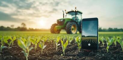 A smartphone stands upright in the soil of a farm field, while a tractor is seen in the background during sunset. This setting highlights the integration of technology in modern agriculture. photo
