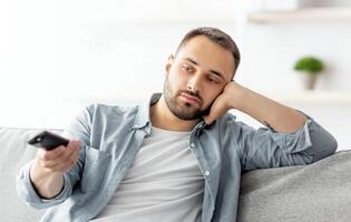 Bored young guy with remote control watching TV on sofa at home, copy space. Millennial Caucasian man switching channels on television, not interested in movie or program photo
