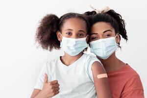 Black girl with adhesive medical band on her shoulder showing thumb up after immunization against coronavirus, sitting on her mother lap, wearing protective face masks, isolated, copy space photo