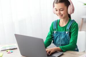 E-learning concept. Smiling female teenager studying online on laptop computer at home. Happy youth using PC to make home assignment, typing on keyboard, chatting with friends or doing homework photo