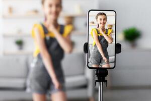 Young Blogger Concept. Cheerful teen girl dancing at camera filming using phone on tripod at home, creating her trendy content on a mobile app to share on social media, selective focus on screen photo