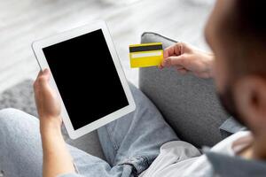 Unrecognizable young guy sitting on couch, holding credit card, shopping online on digital tablet at home, mockup for website or app. Millennial man using web banking service on touch pad photo