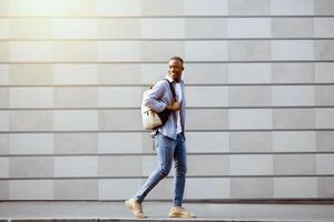 Young black man in casual wear with backpack walking near brick wall on city street photo