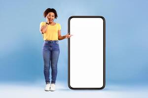 Emotional African American woman cannot believe great offer, pointing at big smartphone with space for new mobile app on screen, blue studio background. Mockup for website. Cellphone display template photo