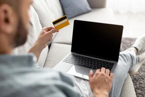 Unrecognizable young guy sitting on sofa, using credit card for online shopping on laptop, space for website mockup. Closeup view of millennial man using web banking service photo