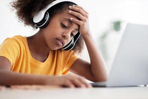 Exhausted black school girl with wireless headset sitting in front of laptop, touching her head, feeling bored or tired while home schooling, need for rest, dreaming about time with her friends photo