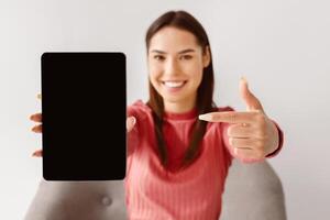 Happy Caucasian woman pointing at tablet pc with black blank screen for mockup, presenting new application, offering space for website, advertising your product or service, selective focus photo
