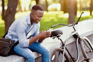 Joyful black man with modern bicycle using cellphone at urban park, copy space. Happy African American biker chatting online with friend, checking mail, browsing internet, playing game outdoors photo