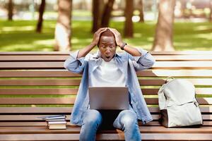 Terrified black man looking at laptop screen and grasping his head in panic on bench at park. African American student finding mistake in his project, failing to meet term paper deadline photo