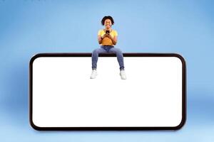 Cheerful young African American lady sitting on big smartphone with empty screen, using mobile device, demonstrating mockup for website, advertisement or app over blue studio background photo