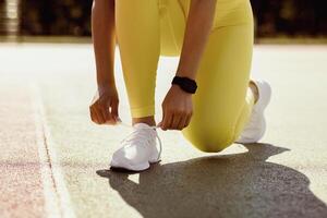 Training Concept. Closeup cropped view of unrecognizable black female athlete in bright yellow leggings tying her shoelaces before jogging outdoors in the morning, wearing white sneakers photo