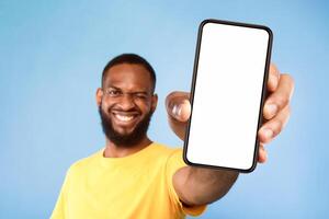 Mobile app advertisement. Smiling young African American man showing smartphone with empty screen, winking on blue studio background, selective focus. Cellphone display mockup, space for ad or website photo
