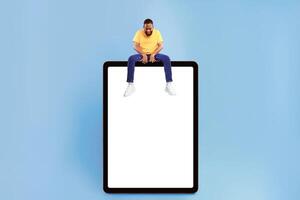 Attractive young African American guy sitting on top of giant tablet computer, looking down at empty screen on blue background, mockup for app design. Touch pad website template, cool online store ad photo