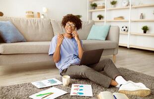 Happy black teenager sitting on floor with laptop and study materials, speaking on smartphone at home. Remote education, distance communication during covid lockdown concept photo