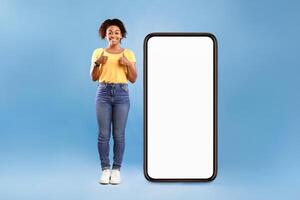 Best mobile offer. Smiling African American lady demonstrating giant smartphone with white screen and showing thumbs up with both hands on blue background, mockup copy space for app or website design photo