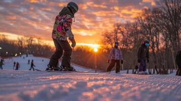 Young skiers practice their skills on a snowy slope as the sun sets behind them, casting warm colors across the winter sky while parents watch from a distance. photo
