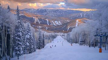 Skiers navigate down a snowy slope surrounded by tall trees, with majestic mountains in the background illuminated by a colorful sunset, creating a serene winter atmosphere. photo