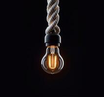 Glowing light bulb suspended from the top of dark background by thick, twisted white rope photo