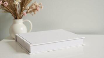 A white book on a table next to a vase of flowers photo