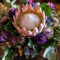 In a sunlit setting, a striking king protea showcases its unique petals, complemented by vivid purple thistles and rich greenery. Nature's artistry shines through in this captivating bouquet photo