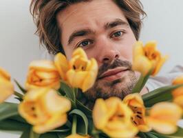 A young man with a gentle expression admires a vibrant bouquet of yellow tulips, surrounded by their lush green leaves. The warm colors evoke a sense of calm and joy in a cozy indoor setting photo