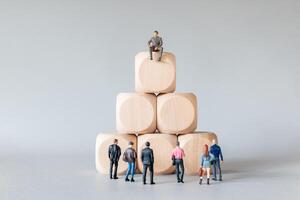 Miniature business stand at the base of a pyramid made of wooden blocks, looking up at a leader sitting on top. photo