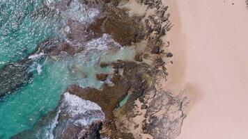 Aerial view of a beach in Oahu, Hawaii, with turquoise waves crashing on rocky outcrops, soft sandy shores, and lush greenery framing the scene. video