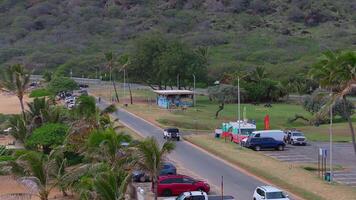 Aerial view of a parking lot near a green hillside on Oahu Island, Hawaii, with a mural covered building, palm trees, and a curving road in motion. video