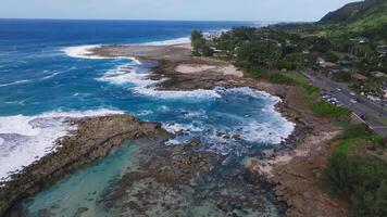 Aerial view of a rocky coastline on Oahu island, Hawaii, with waves crashing, a small beach, lush greenery, and a residential area with a coastal road. video