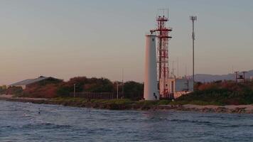 A white lighthouse with a red structure stands near a rocky shoreline as gentle waves move. Palm trees, industrial structures, and mountains are visible. video