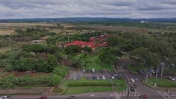 Aerial drone view of the Dole Pineapple Plantation with red roofed buildings, pineapple fields, parking areas, and rolling hills under a cloudy sky. video