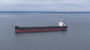 Aerial view of a stationary cargo ship with a red hull and white superstructure in calm waters near Seattle, with forested hills in the background. video