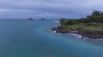 Serene aerial view of Oahu's coastline, featuring Mokulua Islands, beachfront homes, lush greenery, rocky outcrops, and turquoise ocean under overcast skies. video
